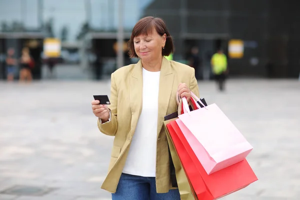 Smiling stylish middle aged woman with credit, debit card and shopping bags standing on shopping mall background. Autumn holidays, discounts, black Friday. concept of consumerism, sale, rich life.