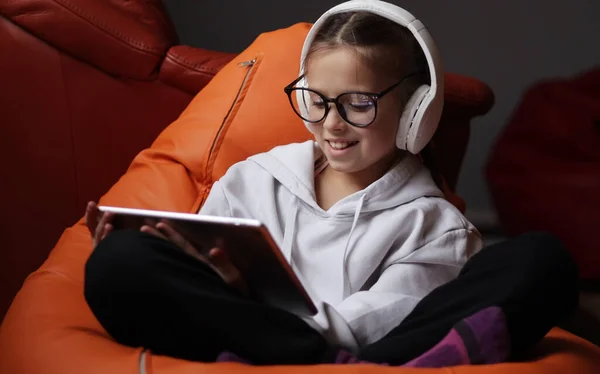 stock image Cute smiling teen girl in glasses and headphones is holding digital tablet talking with remote teacher tutor on social distance video conference call elearning online virtual class at home. Children