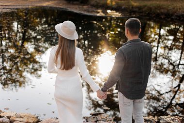 Sonbahar parkında göletin kenarında el ele tutuşan şık, genç bir çiftin arka görüntüsü. Beyaz elbiseli, şapkalı ve şapkalı bir kadın Sevgililer Günü 'nde biriyle çıkar. Mutlu aile aşk konsepti.