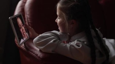 Çevrimiçi sanal aile sohbet videosu toplantısı sırasında dijital tabletle dijital tablet üzerinden karanlık oturma odasındaki kanepede uzanan görüntülü konuşma sırasında arkadaşıyla, kız kardeşiyle, annesiyle ya da öğretmeniyle konuşan gülümseyen bir genç kız..