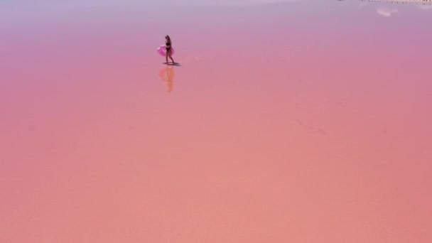 空中展望 黒水着の若い女性は 彼女の手の中にインフレータブルピンクのフロートを運ぶピンクの塩湖で楽しいダンスを持っています — ストック動画