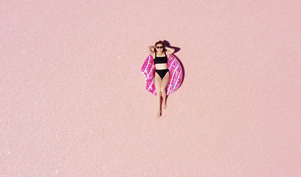 World Tourism Day. Above view of young happy woman in sunglasses and black bikini sunbathing lying on an inflatable pink donut in a sunny pink lake. Enjoying holiday vacations. Place for advertising.
