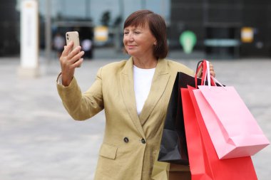 Alışveriş çantaları ve akıllı telefonları olan şık yetişkin kadın blogcu alışveriş merkezi yakınlarındaki sosyal ağlarda takipçilerle iletişim kuruyor. Tüketim, satış, zengin hayat, sanal hayat kavramı. Kara Cuma.