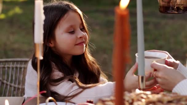 幸せな家族の日 お母さんの手による白いカップから 愛らしい子供の女の子が熱いお茶を飲みます 娘と一緒にお母さんとお父さんでピクニック感謝祭のテーブルで燃えるキャンドルと秋の公園でパイ — ストック動画