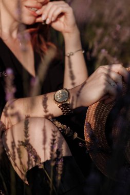 Siyah elbiseli, tanınmayacak kadar seksi bir kadın, elleriyle dudaklarına dokunuyor ve lanet lavanta tarlasında oturuyor. Elindeki altın saate ve hasır şapkaya odaklan. Doğal güzellik konsepti. Kadınlar Günü.