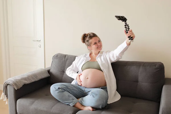 在现代公寓的沙发上 妈妈和流行的维基百科正在为在线博客录制视频 并在智能手机相机上展示她赤身裸体的大肚子 怀孕与博客概念 — 图库照片