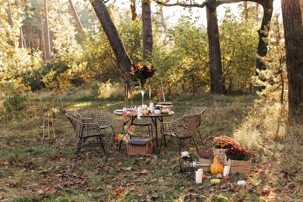 Cozy autumn picnic in the park. Close up of table setting with white plates, cutlery, glass vase with colourful wildflowers, homemade apple pie, maple leaves, burning candles on wooden table outdoors