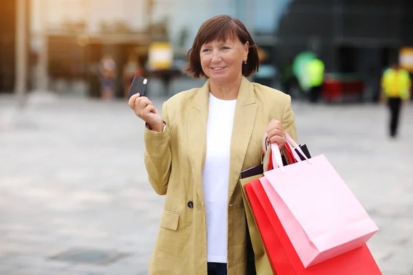 stock image Smiling stylish middle aged woman with credit, debit card and shopping bags standing on shopping mall background. Autumn holidays, discounts, black Friday. concept of consumerism, sale, rich life.