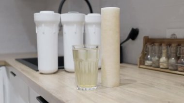 Mutfaktaki ahşap masada su filtresi ve bir bardak paslı su kullanılmış. Üç filtre kartuş ile plastik set ters osmoz su arıtma sistemi içilebilir durumda..