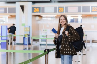 Seyahat, tatil, turizm. Havaalanı terminaline giriş yaptıktan sonra pasaportunu gösteren, kameraya gülümseyen, sırt çantası taşıyan mutlu ve çekici genç bir kadın turist..