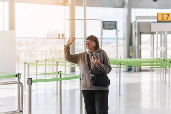 幸せな笑顔の成熟した旅行者の女性は 休暇で新国への旅行から到着した後 空港ターミナルで彼女に会った誰かを迎えるためにスマートフォンと波の手を握っています 引退旅行 — ストック写真