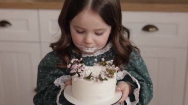 Sevimli küçük kız, süslü bir elbise giyer bir doğum günü partisinde çiçekten bir ısırık alır. Mutlu çocuk, pis suratından beyaz krema yalar ve kapalı mekanda başparmağını kaldırır..