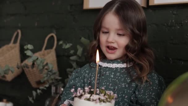 愛らしい小さな女の子4 5年は 花で飾られた白い誕生日ケーキを保持し 燃えるキャンドルを吹こうとしています 子供はお祝いのスタイリッシュなレトロドレスを身に着けています準備ができています彼女の誕生日パーティーに自宅 — ストック動画