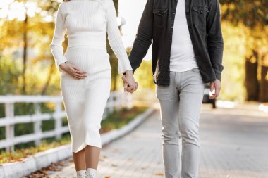 Hamilelik ve Ebeveynlik kavramı. Adam sonbahar parkında hamile karısıyla yürüyor. Mutlu stil sahibi genç aile güneşli bir günde hafta sonu dışarıda dinleniyor.