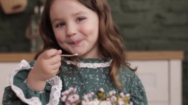 Doğum günü partisi. 4-5 yıllık mutlu küçük kız çocuğu zevkli elbiseler giyiyor. Zevkle yemek yiyor. Tahta kaşık, süslü çiçekli doğum günü pastası..