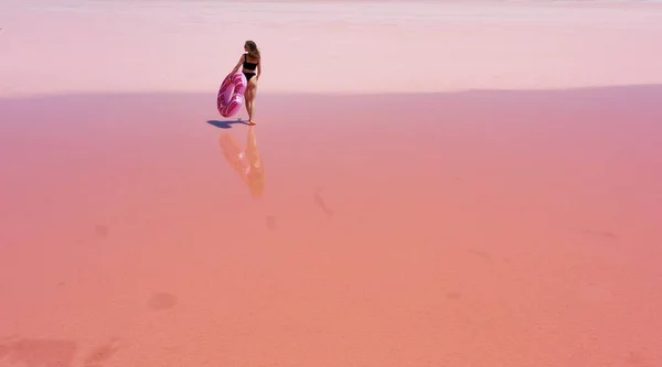 空中风景 穿着黑色泳衣的年轻女子手里拿着一个充气粉色的花环 在粉色的盐湖边跳舞 玩得很开心 — 图库照片