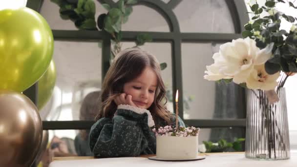 子供の誕生日パーティー 愛らしい少女は 誕生日ケーキ装飾された花と燃えるキャンドルでテーブルに座って 願い事をします 風船のある部屋でお祝いのドレスの子供 花瓶の花の花束 — ストック動画
