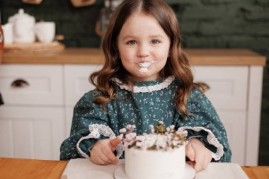 Sevimli küçük kız, süslü bir elbise giyer bir doğum günü partisinde çiçekten bir ısırık alır. Mutlu çocuk, pis suratından beyaz krema yalar ve kapalı mekanda başparmağını kaldırır..