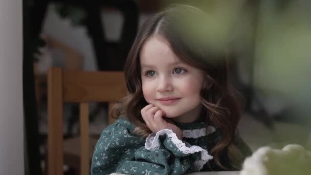 可爱的小女孩 穿着节日的乡村服装 面带微笑 看着相机 坐在桌旁 双手托着下巴 待在家里 快乐童年的概念 儿童节 — 图库视频影像