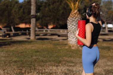 Spor giyim ve kulaklık takan sportif atletik genç bir kadın, palmiye ağaçlarının arka planında köpük masaj silindiriyle ayakta duruyor. Dünya Sağlık Günü. Ekipmanlı spor ve spor, açık havada dolaşın.