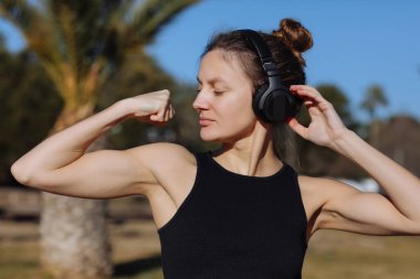 Spor kıyafetli genç bir kadın kaslarını, pazılarını göstermek için kollarını kaldırıyor, güçlü elini gösteriyor, açık havada zayıf figürlere sahip, zaferden emin, bağımsız görünüyor, gülümsüyor..