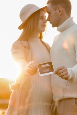Unrecognised of a pregnant woman with her husband are holding an ultrasound scan photo of the unborn child outdoors on nature in sunlights. Happy motherhood and parenthood concept.