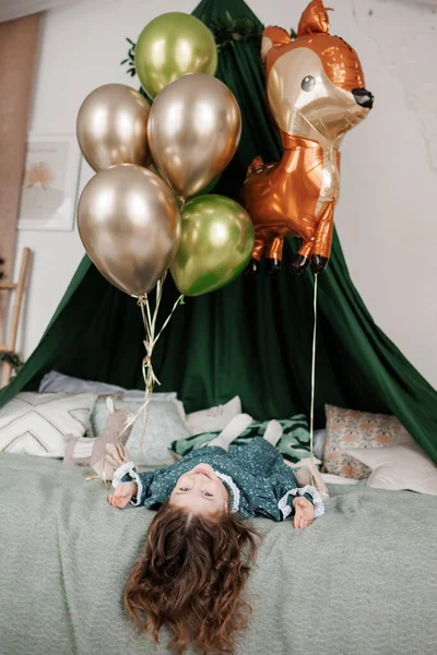 子供の誕生日のお祝い 幸せな女の子のトップビュー4 5年スタイリッシュな素朴なスタイルのドレスを着てベッドの上に横たわっている 笑顔とカメラを見て 風船で誕生日パーティーのために装飾されたベッドルーム — ストック写真