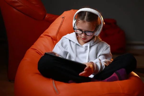 stock image Concentrated teen girl in headphones plays game, communicate on internet social media, listens to music or online studies at home on digital tablet sitting on sofa. Children tech addiction concept.