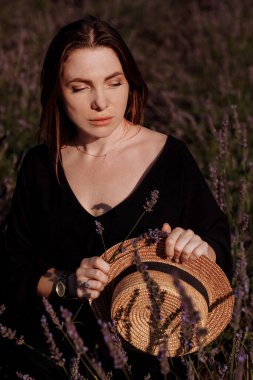 Siyah elbiseli genç seksi kadının portresi çiçek açan mor lavanta tarlasında oturup kameraya bakıyor. Çekici esmer kız hayattan zevk alıyor ve hayal kuruyor. Doğal güzellik konsepti.