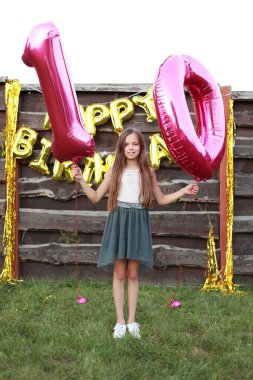 Şirin küçük kız iki pembe folyo balon tutuyor, açık havada 10 haneli rakamlar. Altın balonlarla süslenmiş ahşap duvar. Doğum günün kutlu olsun. 10. yıl dönümü kutlaması..