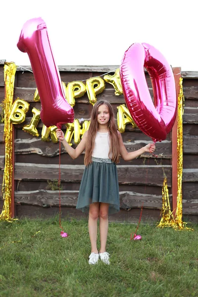 Cute little girl holds two pink foil balloons, digit 10 outdoors. Wooden wall decorated with golden balloons inscription happy birthday.Ten years anniversary celebration event, party at home backyard.