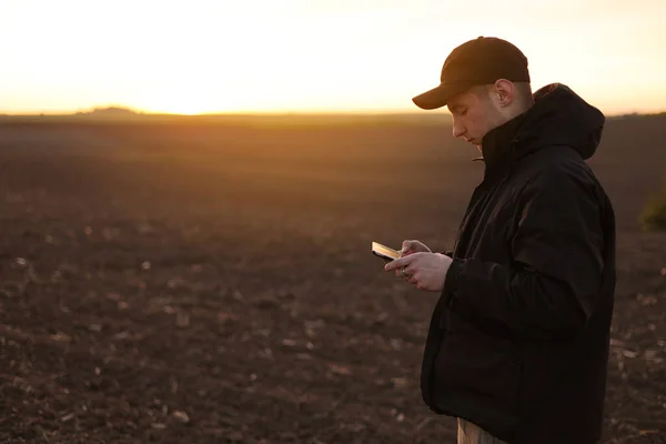 Smart farming technology and agriculture. Farmer uses digital tablet on field with plowed soil at sunset. Checking and control of soil quality, land readiness for sowing crops and planting vegetables.