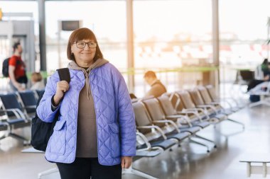 Orta yaşlı gözlüklü, el çantalı, sırt çantalı bir kadın havaalanının kapısında elinde akıllı bir telefonla bekliyor. Aktif macera, hayat tarzı seyahatini emekliye ayırdı