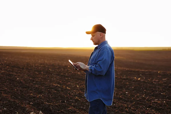 Smart farming technology and agriculture. Farmer uses digital tablet on field with plowed soil at sunset. Checking and control of soil quality, land readiness for sowing crops and planting vegetables.