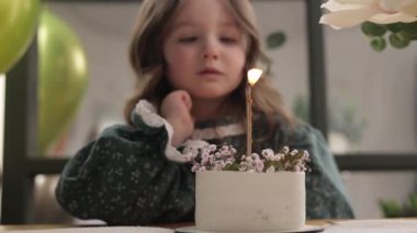 Komik küçük kız, çiçeklerle süslenmiş doğum günü pastasına yanan mum üflemeye çalışıyor. Mutlu çocuk şık bir elbise giyer ve doğum gününü dekore edilmiş oturma odasında kutlar..