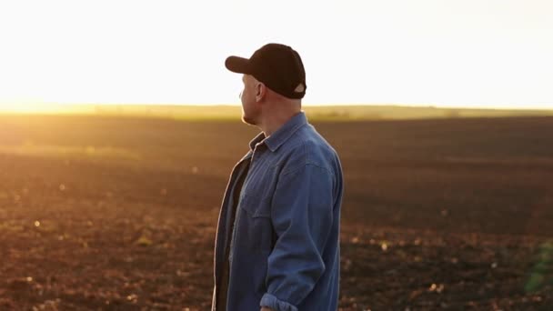 Side view of male farmer standing and looking in cultivated plowed field at sunset in spring. Owner agricultural farm is checking and examining farmland before sowing agriculture crops. Agribusiness