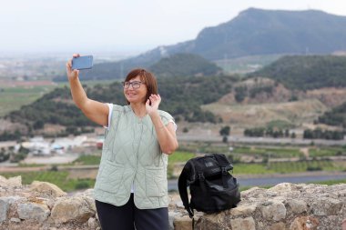 Yetişkin gezgin kadın blogcu, sırt çantasıyla tepenin üstünde duruyor. Akıllı telefonu tutuyor, takipçileriyle iletişim kuruyor ve sosyal medyada dağları gösteriyor. Avrupa 'da tek başına güvenlikli kadın sırt çantası.