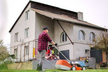 Genç adam evinin arka bahçesindeki çim biçme makinesiyle çimleri biçiyor. Baharın bulutlu akşamında koca bahçeyle ilgilenir. Modern elektrikli kablosuz bahçe ekipmanları. Peyzaj işi konsepti.