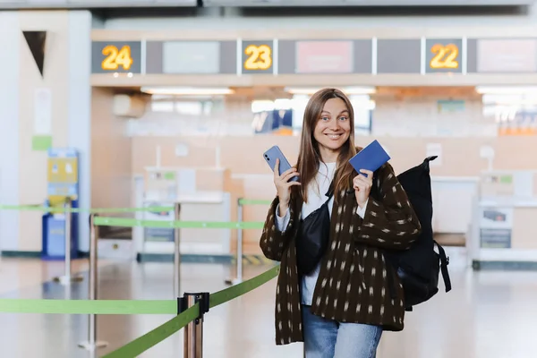 観光の概念 航空会社のサービスデスクでチェックインした後に公共空港ターミナルでパスポートを示す幸せな魅力的な若い女性観光客 カメラで笑顔 バックパックを運ぶ — ストック写真