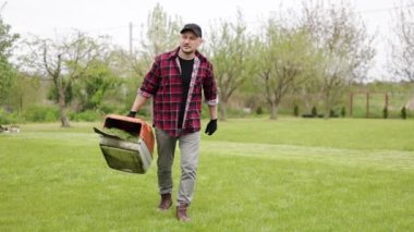 Günlük giysiler içindeki bahçıvan bahar günü evin arka bahçesinde modern elektrikli çim biçme makinesi kesilmiş plastik çim biçme makinesi kutusu taşıyor. Gübre veya sebze bahçesi için çim kesme makası kullanılıyor.