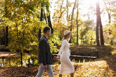 Çift, erkek ve hamile eş gün batımında el ele tutuşup sonbahar parkında yürüyorlar. Mutlu aile doğada hamileliğin tadını çıkarıyor. Anneler Günü ve Kadınlar Günü konsepti