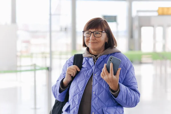 손가방을 안경을 중년의 여성은 스마트폰을 비행기 탑승구 옆에서 기다리고 있었다 — 스톡 사진