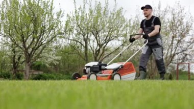 Koruyucu giyimli profesyonel bahçıvan arka bahçedeki modern benzin kablosuz çim biçme makinesini kullanarak yeşil çimen biçiyor. Mevsimlik peyzaj tasarımı. Arkaplanda çiçek açan ağaçlar.