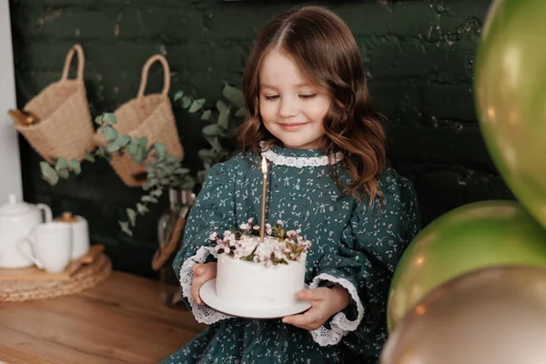 愛らしい小さな女の子4 5年は 花で飾られた白い誕生日ケーキを保持し 燃えるキャンドルを吹こうとしています 子供はお祝いのスタイリッシュなレトロドレスを身に着けています準備ができています彼女の誕生日パーティーに自宅 — ストック写真
