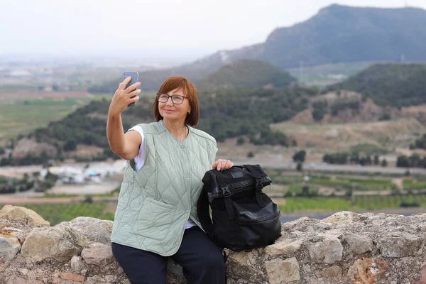 Adult travel woman blogger with backpack stands on hill after hiking up holds smartphone, communicates with her followers and shows mountains at social media. Safety solo female backpacking at Europe.