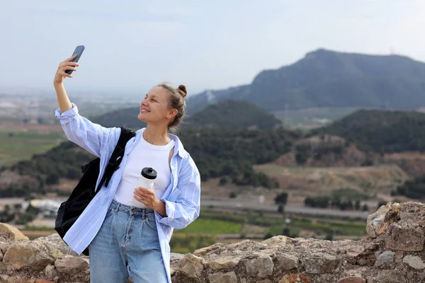 Attractive young travel woman blogger with backpack stands on hill after trekking up holds smartphone, communicates with her followers and shows landscape at social media. Safety solo trip at Europe.