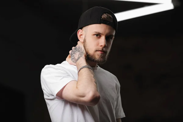 Stylish bearded caucasian young man with tattooed arm in cap and white t shirt looking at camera, light smiling, touching neck with hand on dark background with electric diode lamps. Fashion concept.