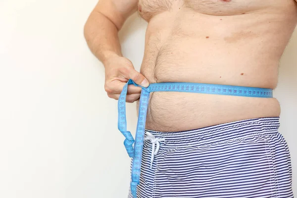 A man measures his fat belly with measuring tape. Concept of weight loss, health problems of obese people. Poor nutrition and sedentary lifestyle. Increased risk of heart illnesses. World Obesity Day.