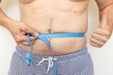 Adam göbeğini mezurayla ölçüyor ve baş parmağını kaldırıyor. Kilo kaybı kavramı, obez insanların sağlık sorunları. Yeme ve aktif yaşam tarzını kontrol etmek. Dünya Obezite Günü.
