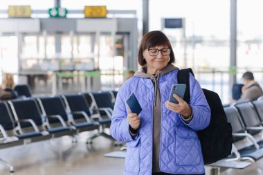 Bagajlı, sırt çantalı ve pasaportlu olgun bir yolcu uçuş saatini kontrol ediyor ya da havaalanı terminalinde akıllı telefondan giriş yapıyor..